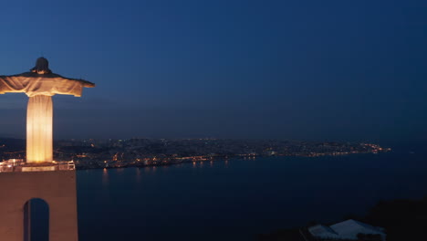 Vista-Aérea-Nocturna-Del-Centro-De-La-Ciudad-De-Lisboa-Con-Luces-En-La-Distancia-Y-Santuario-De-Cristo-Rey-Monumento-En-La-Colina-Que-Domina-La-Ciudad