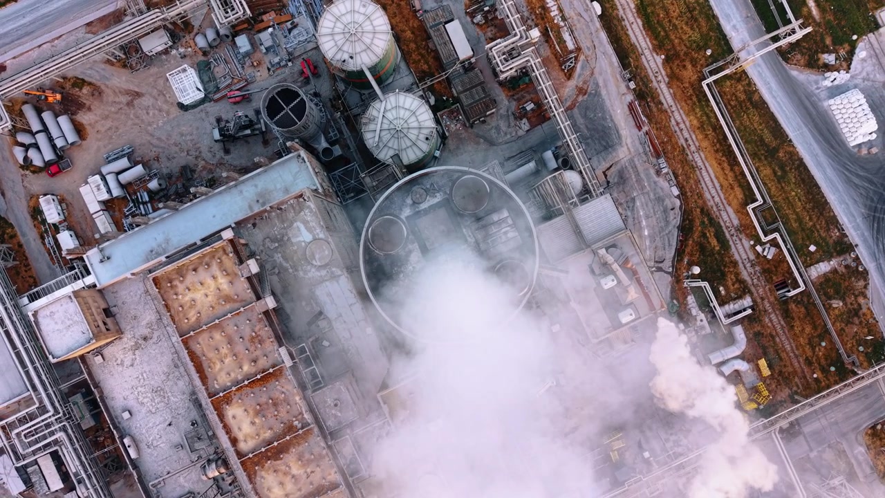 Vista aerea dall'alto di fumo che esce da un edificio di fabbrica. Fumo bianco si sprigiona da uno sfiato circolare sul tetto sopra tubi e strutture in un sito industriale.