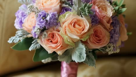 peach and lavender roses, purple lisianthus, dusty miller, and succulents beautifully arranged in a wedding bouquet, displayed on a luxurious sofa