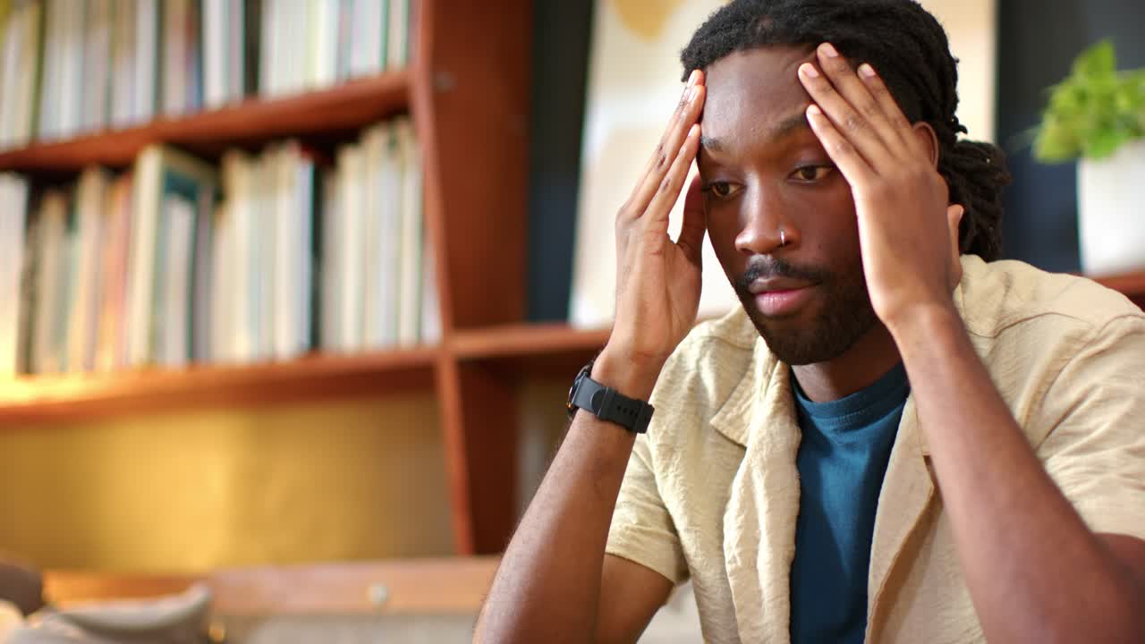 African American man rubbing temples and closing eyes on couch, self-led, bookshelf visible