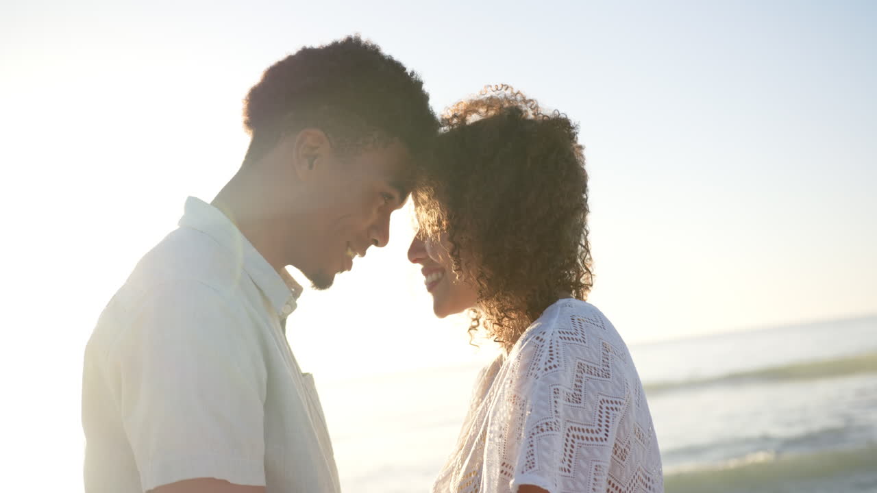 Biracial couple shares a tender moment, foreheads touching at the beach during sunset