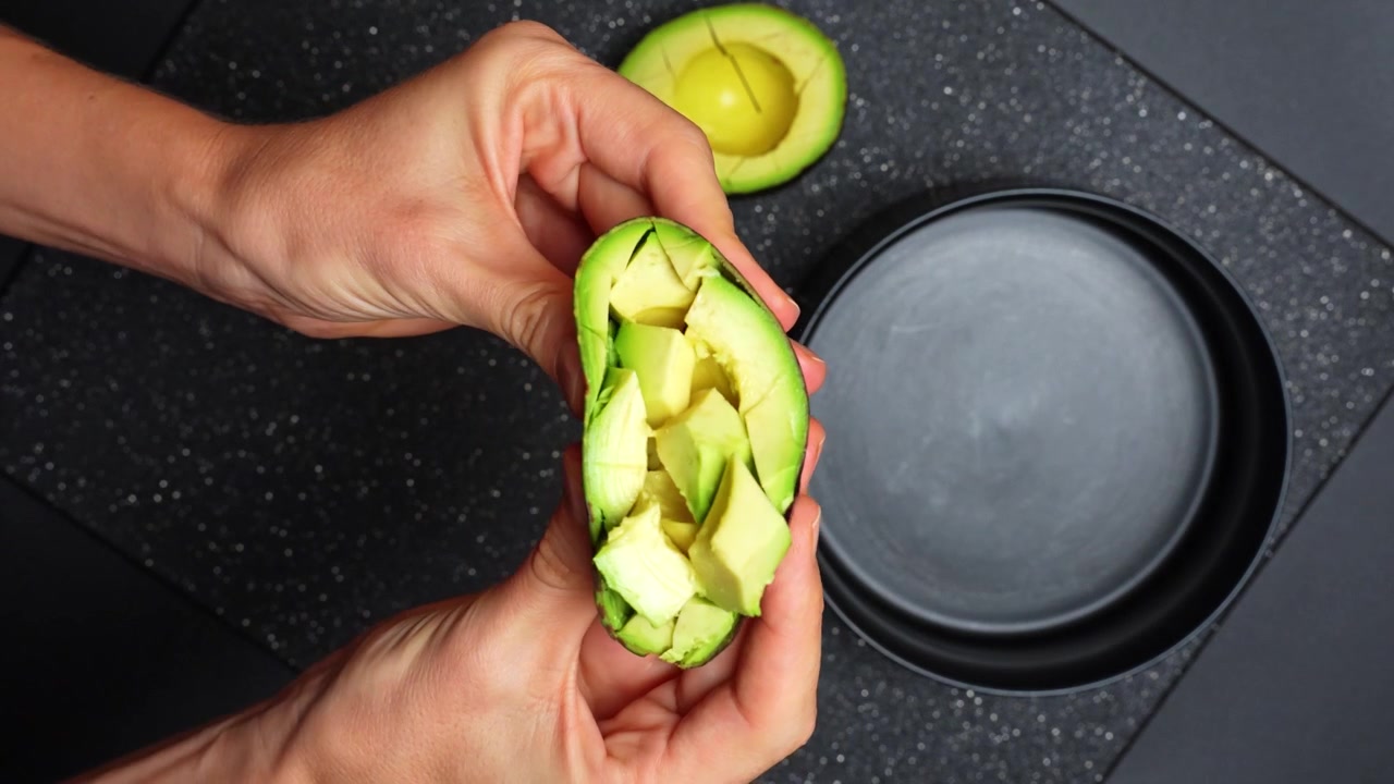Hænder holder halveret avocado med udskåret frugtkød over en skål. Nærbillede af en person, der klemmer en avocadohalvdel for at frigive udskårne frugtstykker ned i en sort beholder.