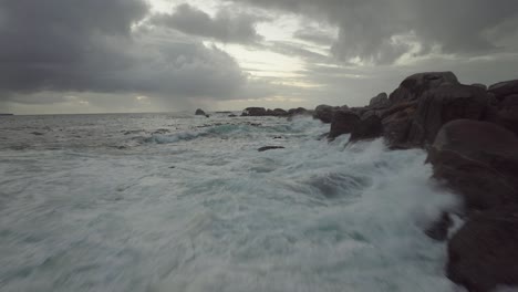 drone volando justo por encima del mar en la playa de camps bay en ciudad del cabo, sudáfrica. está nublado y las olas se están golpeando contra las rocas.