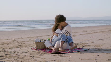 madre sosteniendo y abrazando a su somnolienta hija de rodillas durante un picnic en la playa