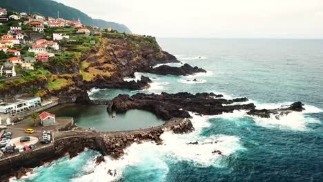 aerial view of the volcanic lava pools of seixal, madeira island, portugal, with crashing waves hitting the rocky coast and some buildings of the village on a cliff in the background