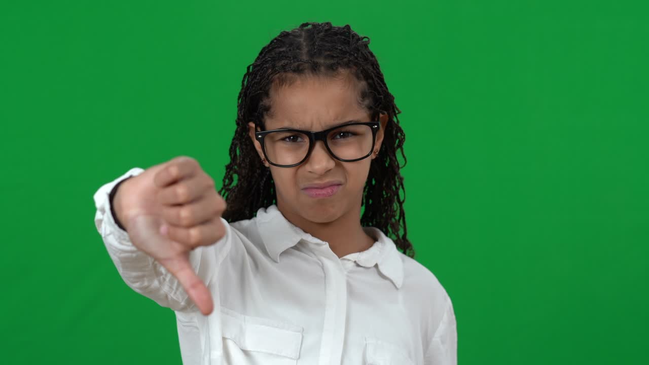Dissatisfied nerd African American teen girl showing thumbs down on green screen looking at camera. Portrait of displeased smart teenager posing at chromakey background. Disapproval concept.