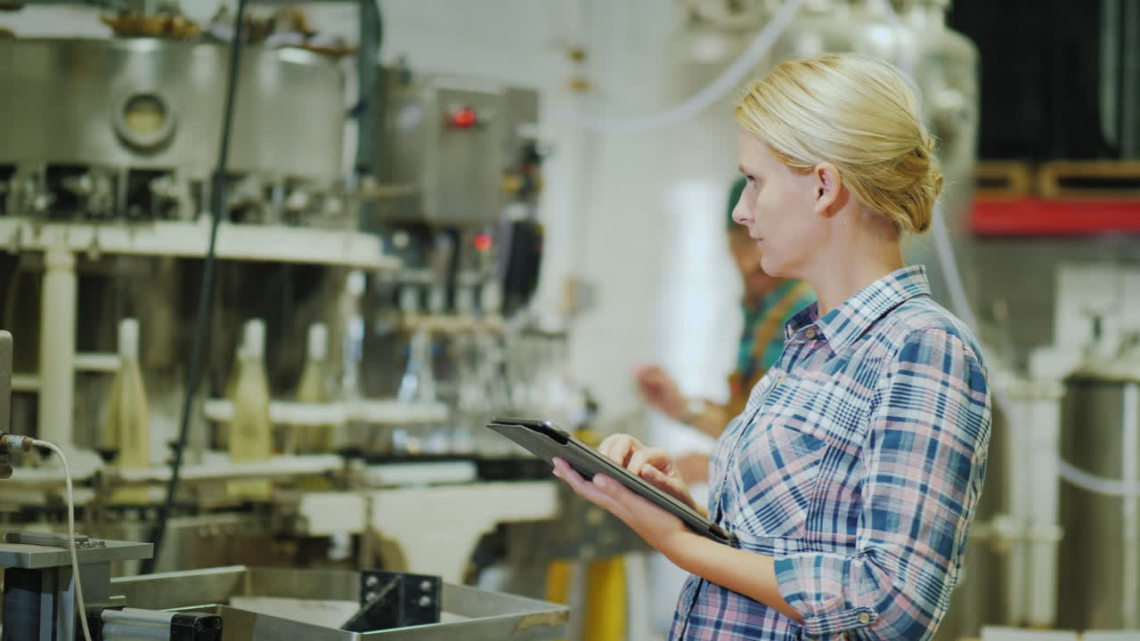 A Woman Is Using A Tablet In A Beverage Factory In The Background Workers Are Working At The Assembl