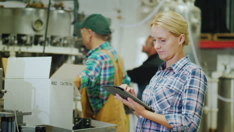 a woman is using a tablet in a beverage factory in the background workers are working at the assembl