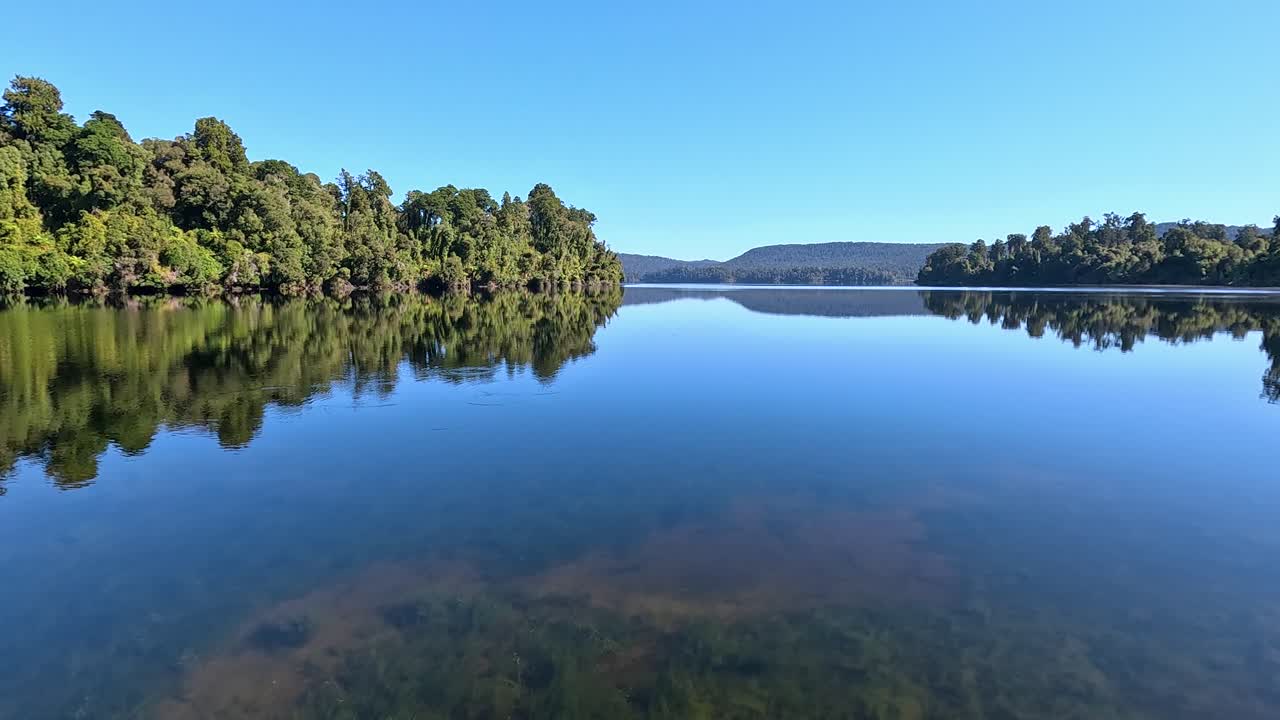 Woman walking along a jetty at Lake Mapourika in New Zealand, South Island. She is walking towards the large open body of water surrounded by dense forest and greenery