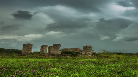 Toma-De-Tiempo-Del-Movimiento-De-La-Nube-Oscura-Sobre-La-Estructura-De-Piedra-Abandonada-Rodeada-De-Praderas-Verdes-Durante-La-Puesta-De-Sol