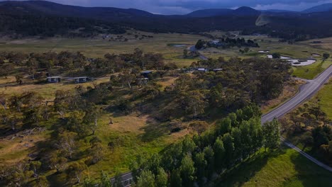 Aerial-shot-of-Crackenback-Alpine-Way-highway-with-farms,-houses-and-cloudy-mountain-ranges,-New-South-Wales,-Australia