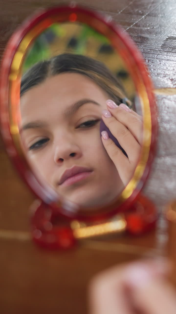 Adolescente reflejada en un espejo rojo, aplicándose una esponja en las mejillas durante una tranquila rutina de belleza con una borla morada en primer plano, reflejos suaves, expresión concentrada, tocador de mesa compacto y delicado