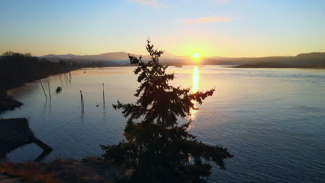 Drone-slightly-panning-to-the-right,-watching-over-a-beautiful-ocean-view-during-sunrise,-surrounded-by-trees-and-mountains