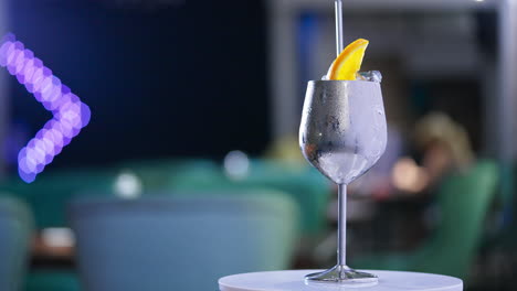 misted metal wineglass with metal straw inside on the rotating table. cocktail with ice cubes and orange slice on top. close up. blurred backdrop.