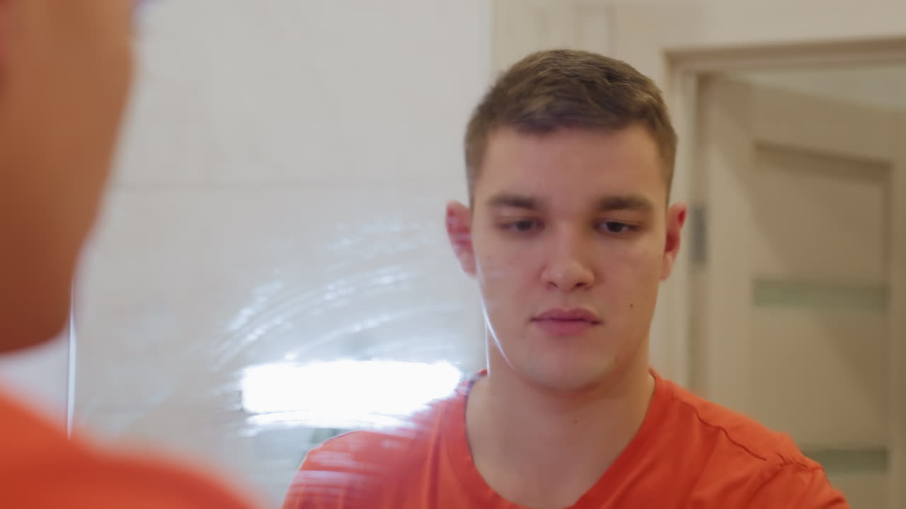 Man in orange shirt spraying soapy water onto bathroom mirror while preparing to wipe surface with soft rag, showing attention to household hygiene and cleanliness