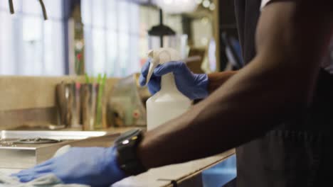 midsection of african american male barista wearing face mask disinfecting bar surface