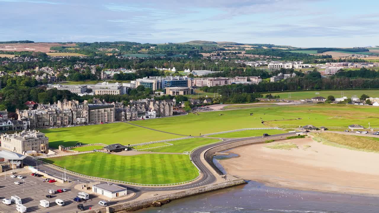 Smooth drone flyover of coastal golf course, historic buildings, parking, and beach in bright daylight