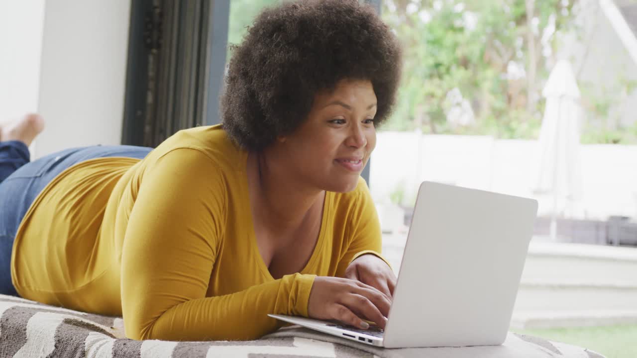 Video of happy plus size african american woman lying on floor and using laptop