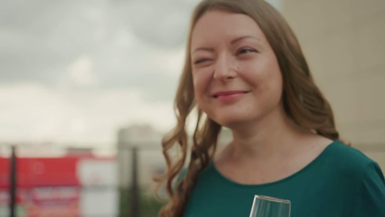 woman holding two wine glasses on terrace, soft smile in green dress, overcast sky and city background, candid portrait capturing gentle anticipation and warm natural light, subtle romantic vibe