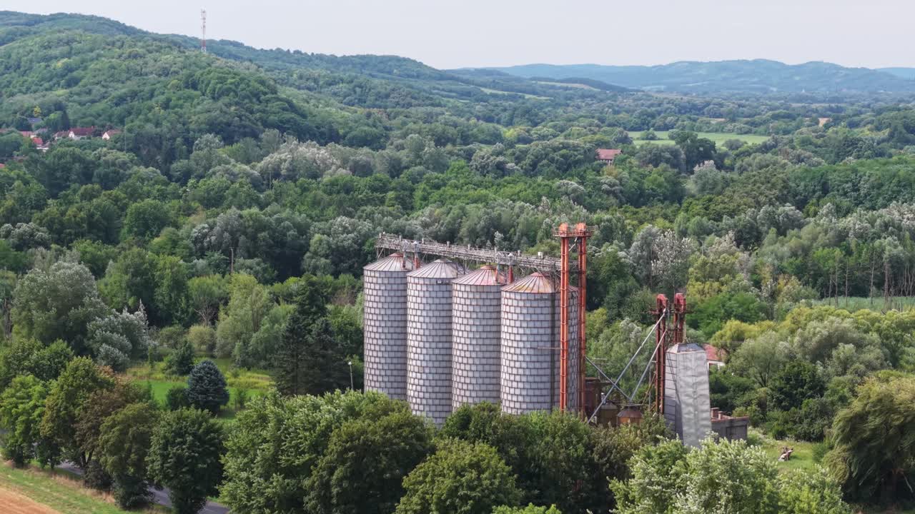 Vier große Getreidesilos mit verrosteten Dächern stehen inmitten eines dichten, grünen Waldes und sanften Hügeln. Die Szene zeigt eine friedliche ländliche Gegend mit landwirtschaftlicher Infrastruktur, umgeben von natürlicher Schönheit.