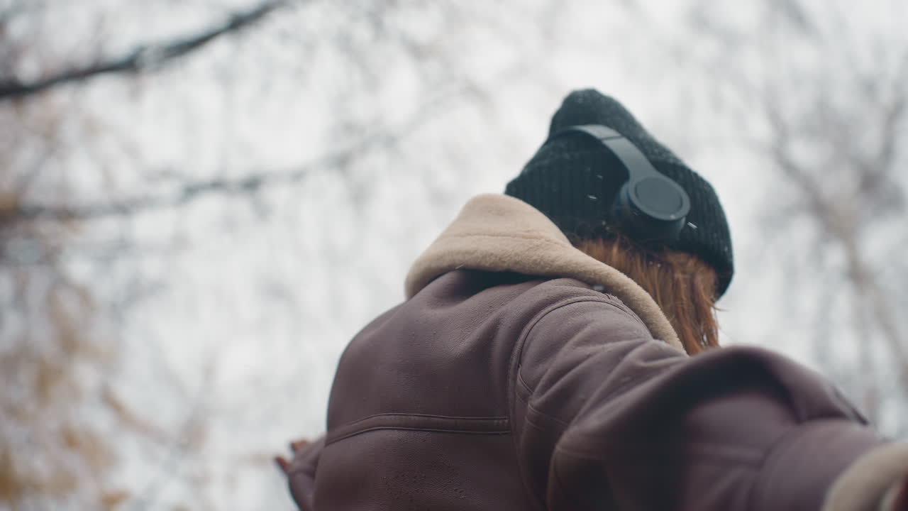 Close shot of lady wearing black knit cap, brown shearling jacket with arms outstretched, walking joyfully outdoors on cool autumn day, enjoying fresh air and nature's beauty
