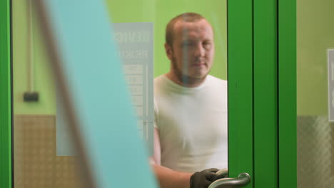 una vista cercana de un hombre saliendo a través de una puerta de vidrio verde con marco verde en polo blanco, con un objeto blanco colocado delante de él