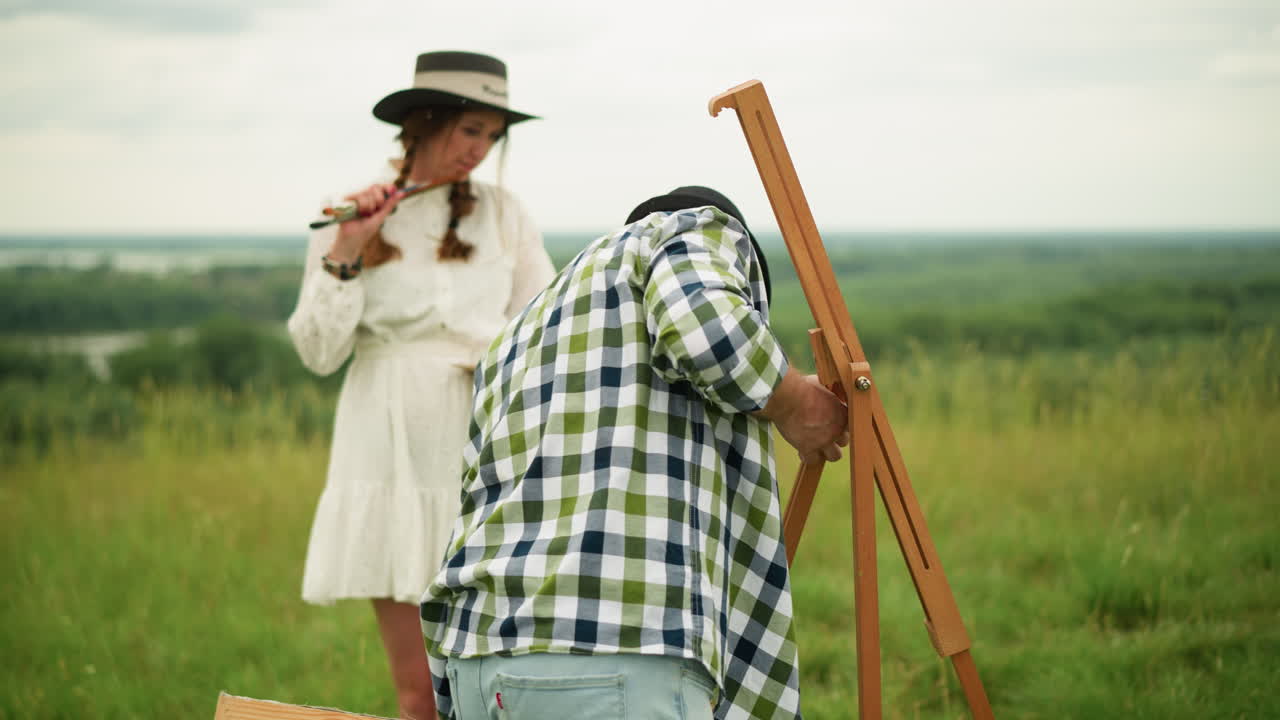 A man wearing a black hat and plaid shirt is focused on setting up a wooden tripod in a grassy field. His partner, in a white dress, stands behind him holding a paintbrush, watching with a smile