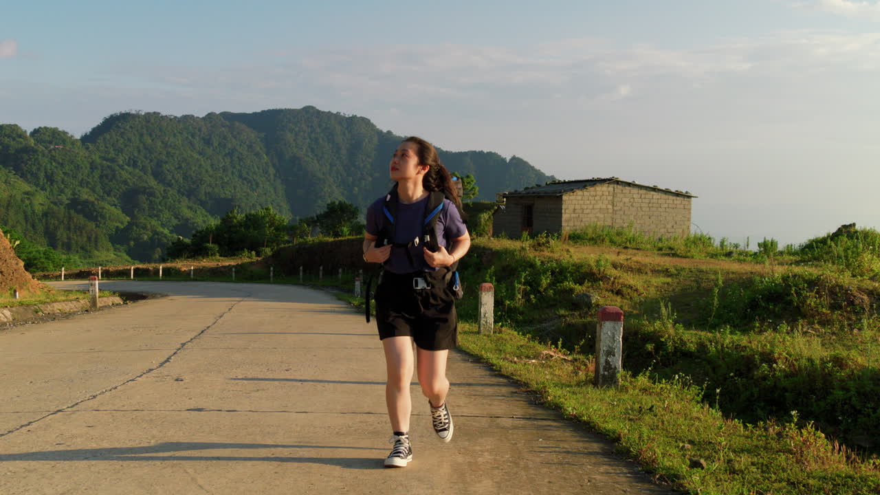 해가 지는 시골 경로를 가로질러 하이킹 배 ⁇ 을 들고 있는 아시아 여성의 전면 추적 전망