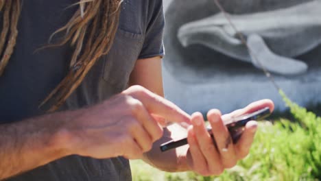 Video-of-midsection-of-caucasian-male-artist-with-dreadlocks-using-smartphone-by-whale-mural-on-wall