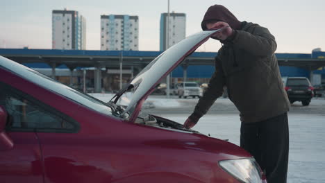 young man in winter jacket removes support rod from raised bonnet of car after engine inspection and prepares to close it, with snowy pavement, parked cars, and city buildings visible in background