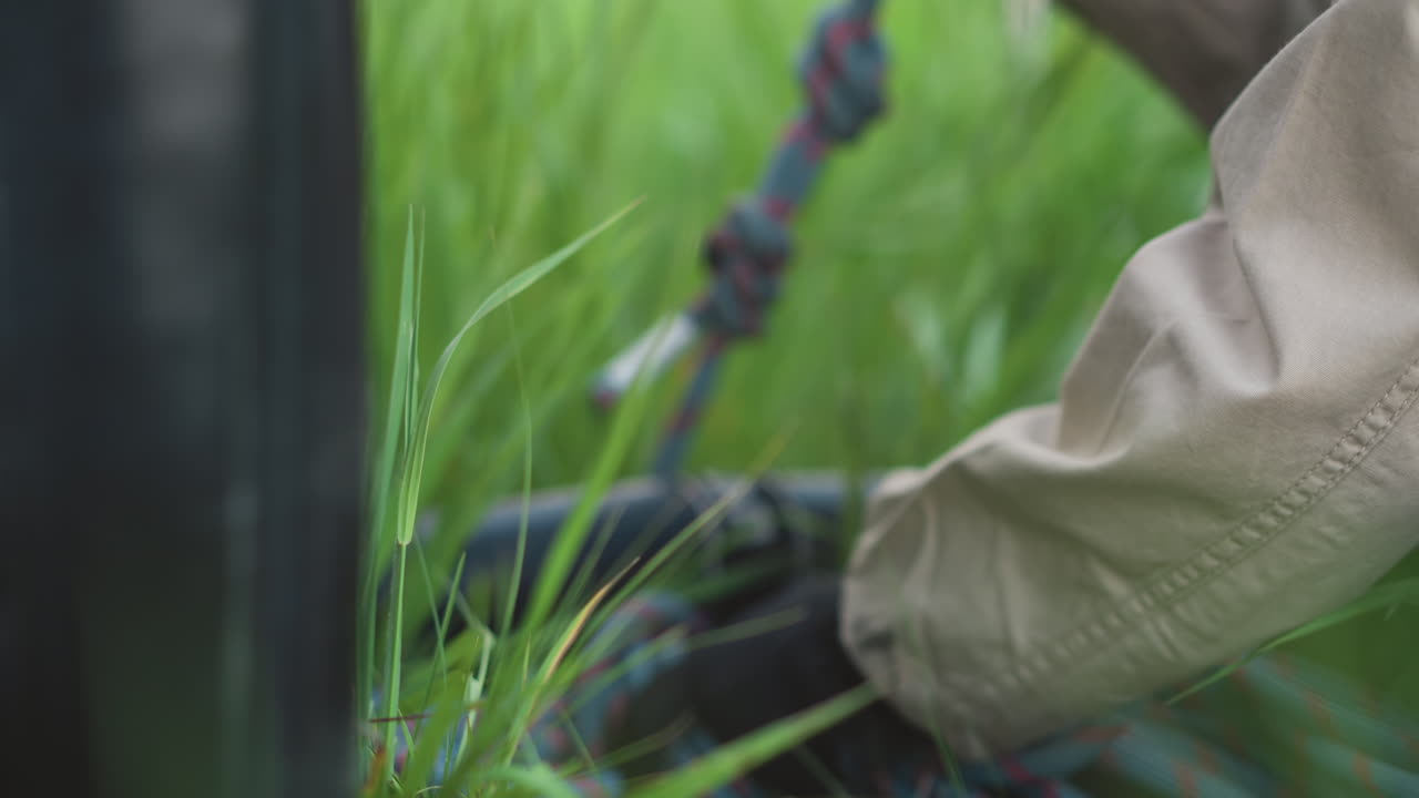 close up of person kneeling in tall grass securing rope and connector under balloon basket frame during preflight setup shot captures focused hands tying safety lines against blurred green field