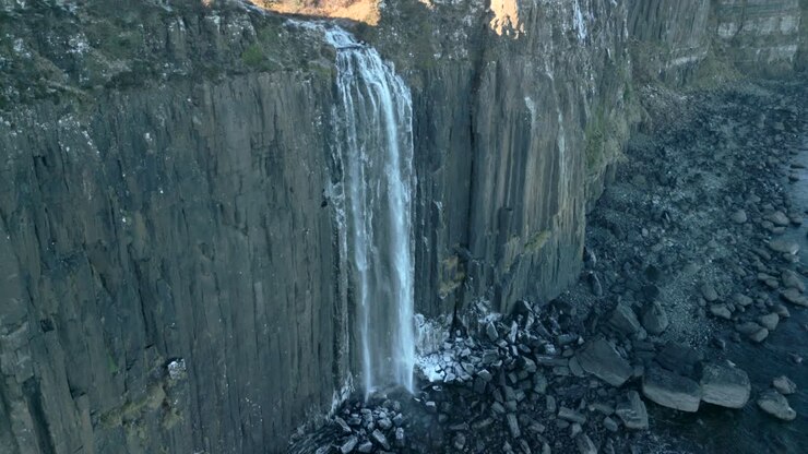 Waterfall half speed slow motion in shadow falling down sheer cliff to frosted rocky beach below in winter at Kilt Rock Waterfall, Isle of Skye, Western Highlands, Scotland, UK