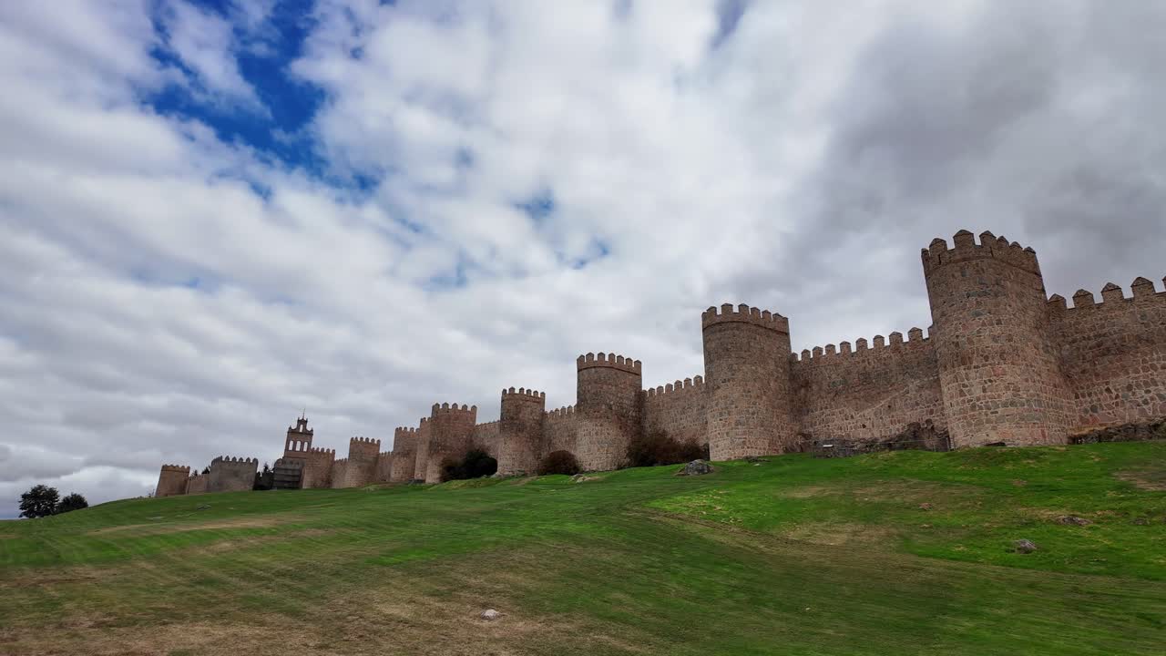 Ampia veduta delle storiche mura medievali di Ávila, Spagna, che si estendono su una collina erbosa sotto un cielo nuvoloso e drammatico, una delle città fortificate meglio conservate d'Europa e un sito patrimonio UNESCO