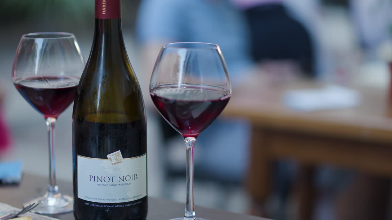 Pinot Noir bottle and glass in focus, blurred people dining outdoors, soft natural lighting