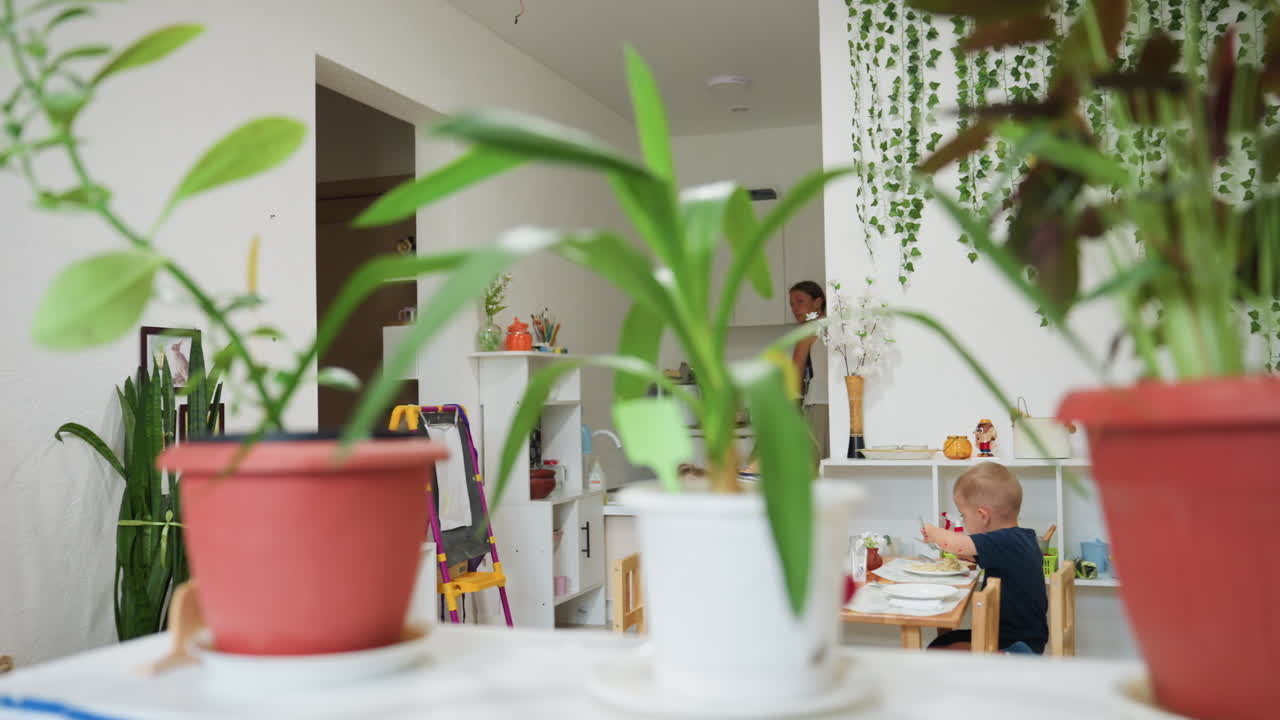Woman assists young children during meal in bright classroom with natural decor and plants. One child in striped clothes stands to return plate while others eat calmly