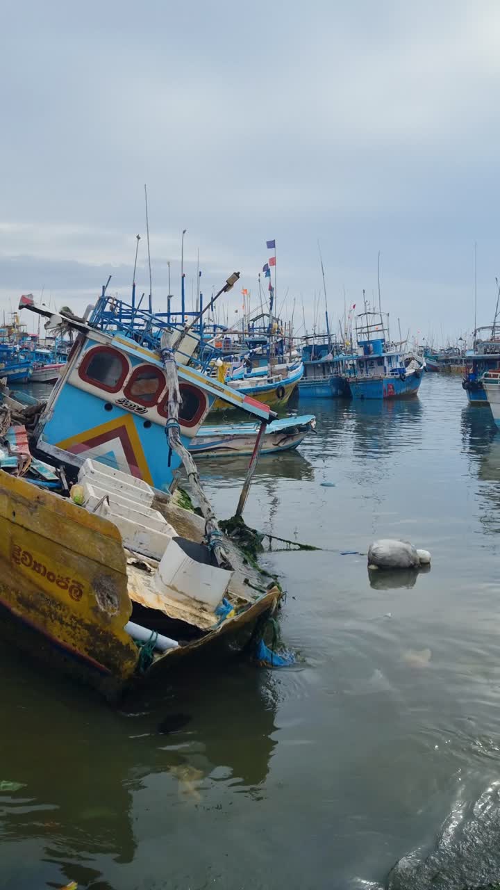 Vertical shot of a damaged, partly sunken boat at Dondra Port fishing market, southern Sri Lanka, with anchored fishing boats in the background on a calm, cloudy monsoon day
