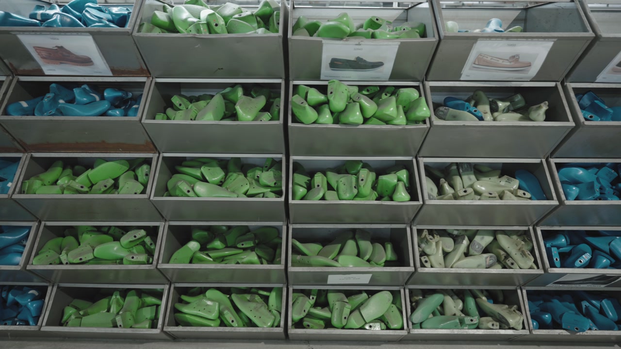 Rows of blue and green plastic shoe molds organised in metal shelf trays in a footwear factory storage room, shoe reference images visible
