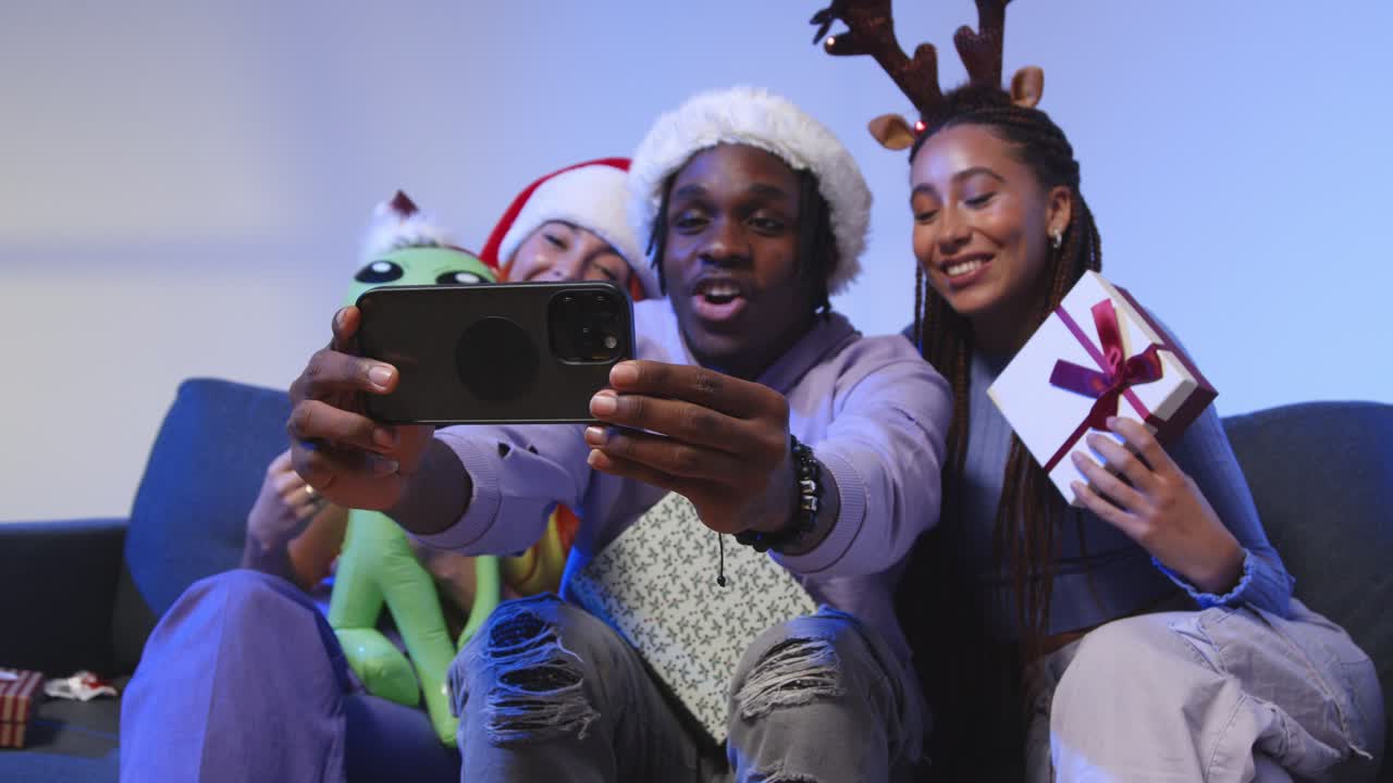 Studio Shot Of Gen Z Friends At Christmas Sitting On Sofa Wearing Santa Hat And Reindeer Antlers Taking Selfie On Mobile Phone 1