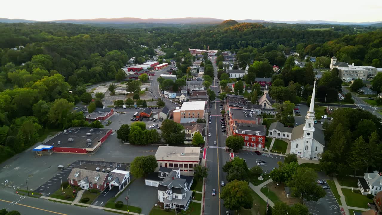 Aerial backwards wide shot of Main Street in Lee, Massachusetts, United States. White church tower and historic buildings in small town. Beautiful autumnal day with colored forest trees in USA