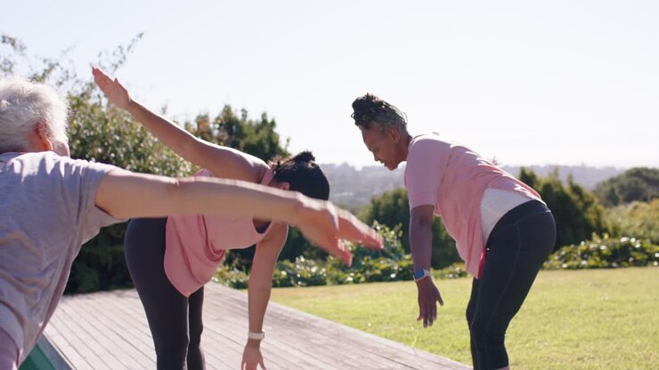 Donne di diverse età e provenienze, inclusa un'anziana, che seguono le istruzioni e fanno stretching su una pedana con tappetini e una bottiglia d'acqua.