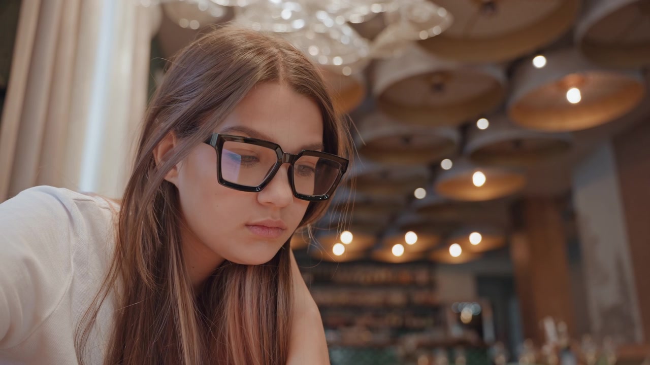 Young woman studying in cafe with glasses, laptop and notebook soft chandelier lighting creates warm bokeh background, pensive expression suggests focus and introspection during evening study session