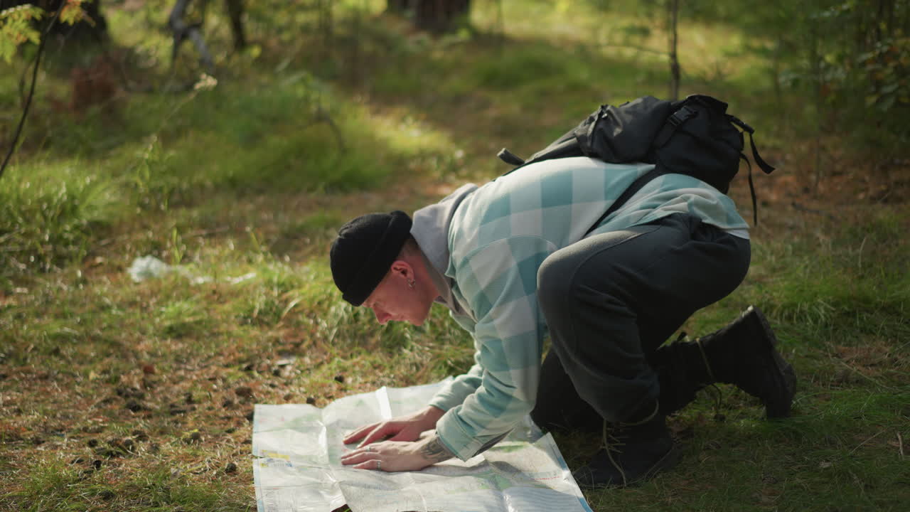 Backpacker kneeling on grass unrolling map and planning route with careful orientation soft light through trees, detailed map spread on ground, focused mapping and decision making
