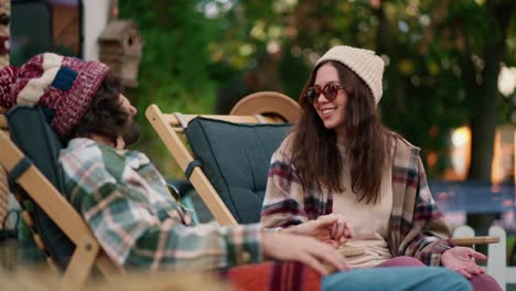 Happy-brunette-girl-in-a-white-hat-with-round-glasses-in-a-plaid-shirt-communicates-with-her-brunette-boyfriend-on-dark-green-chairs-near-a-trailer-in-the-camp-during-her-picnic-outside-the-city-in-the-summer