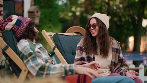 feliz chica morena con un sombrero blanco con gafas redondas en una camisa a cuadros se comunica con su novio moreno en sillas de color verde oscuro cerca de un remolque en el campamento durante su picnic fuera de la ciudad en el verano