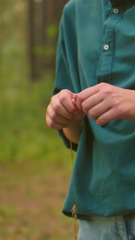 primer plano de una mujer caminando por un sendero forestal, rodeada de árboles altos, sosteniendo una hebra de hierba y cortándola jugosamente, mientras que otra mujer con uñas largas es parcialmente visible