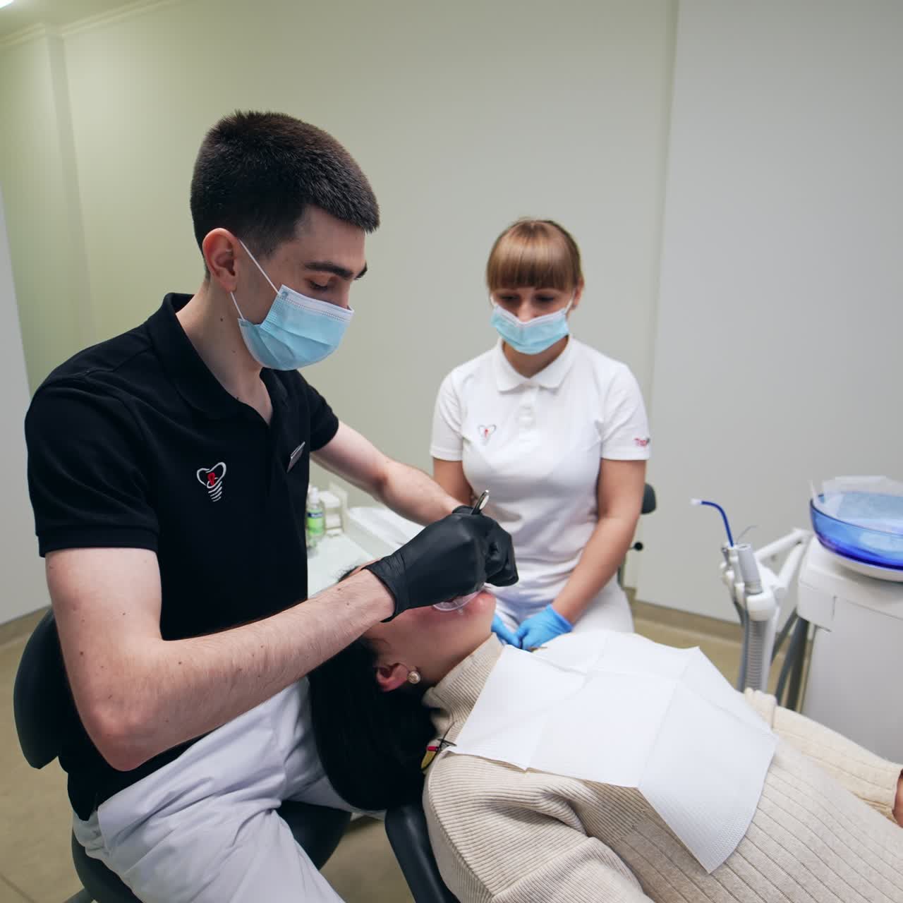 Dentist in black t-shirt working in the light spacious cabinet. Doctor uses metal tool in his work. Female nurse sitting at backdrop