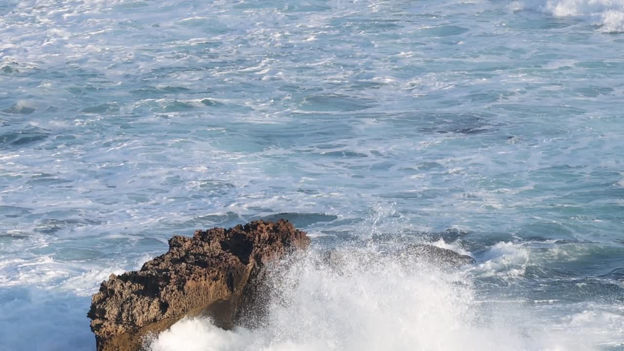 Powerful ocean waves splash and foam against a solitary brown rock in the blue sea