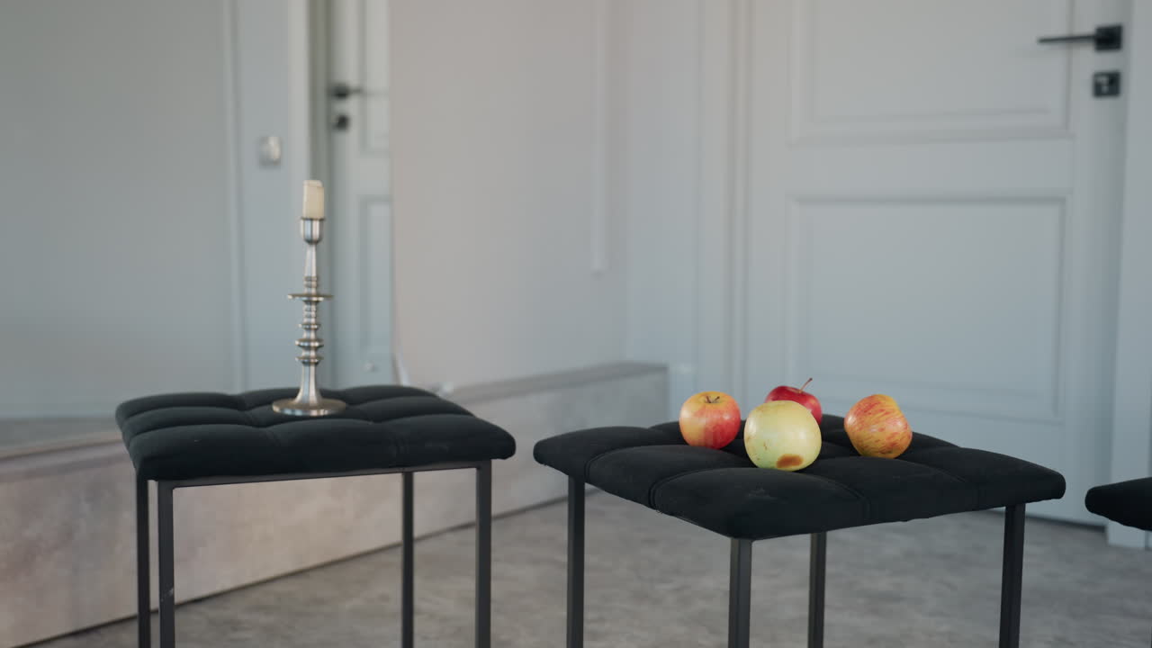 four apples resting on black padded seat next to tall silver candle stand inside bright minimalist studio with mirrored wall and white paneled backdrop highlighting contrast and texture