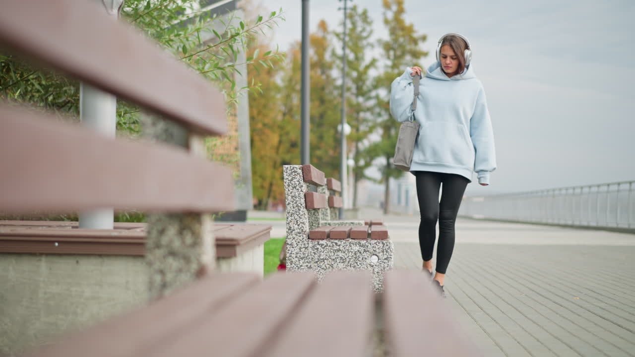 Woman in casual clothes and headphones walking to sit on concrete bench with handbag in background, featuring marcescent trees and street pole in outdoor city park setting