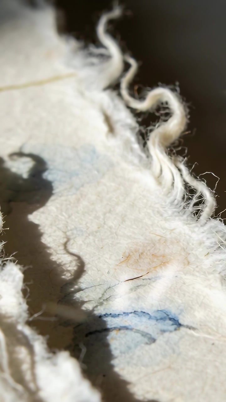 Video verticale: Telecamera che si sposta, rivelando un foglio bianco su un tavolo, evidenziando fibre e una macchia blu.
